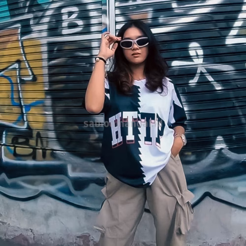 Seorang perempuan bergaya modern mengenakan kaos oversized dengan desain bertuliskan 'HTTP' dan pola warna kontras, dipadukan dengan celana kargo kekinian, berlatar belakang dinding grafiti yang trendi.